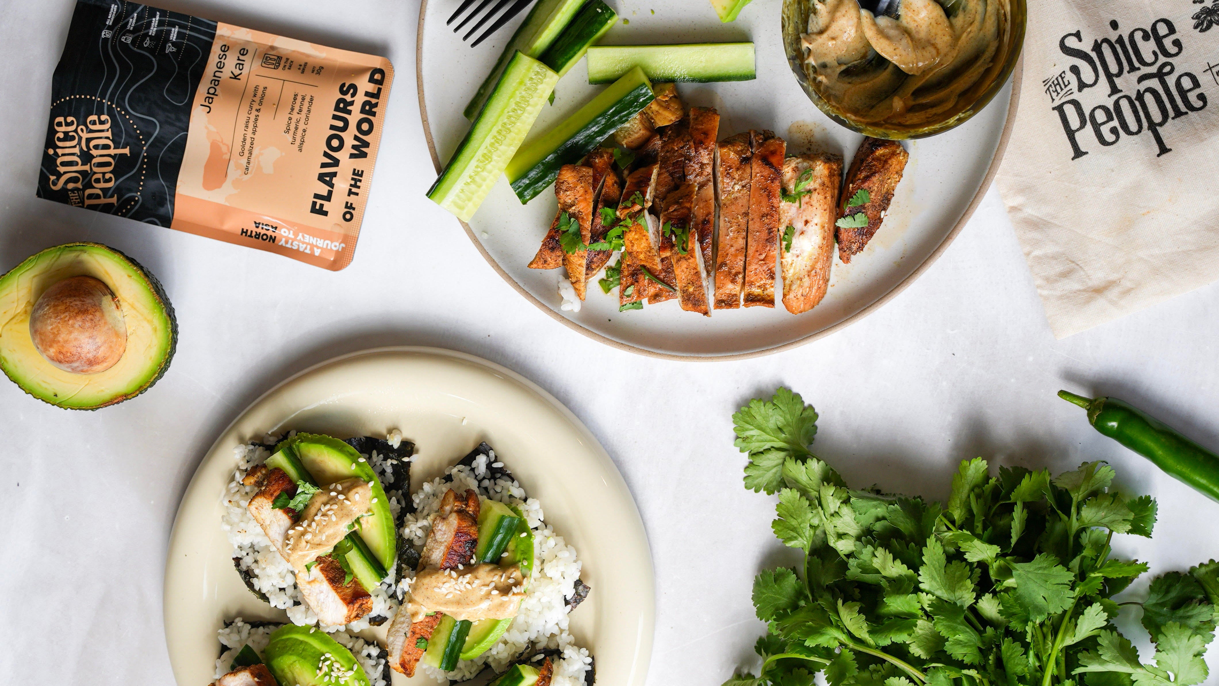 Overhead flat-lay of a plated prepared food dish with sliced grilled chicken, avocado, cucumber, rice, and a small dipping sauce, alongside a Spice People pouch and a branded napkin on a light surface.