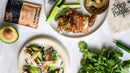 Overhead flat-lay of a plated prepared food dish with sliced grilled chicken, avocado, cucumber, rice, and a small dipping sauce, alongside a Spice People pouch and a branded napkin on a light surface.