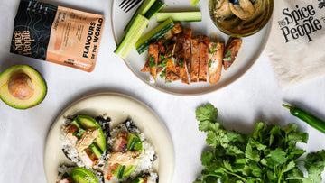 Overhead flat-lay of a plated prepared food dish with sliced grilled chicken, avocado, cucumber, rice, and a small dipping sauce, alongside a Spice People pouch and a branded napkin on a light surface.